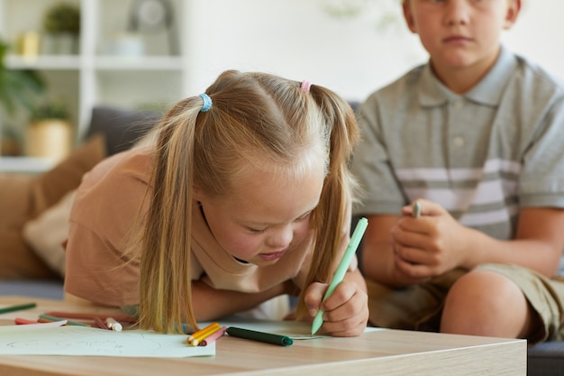 Portrait of cute blonde girl with down syndrome writing or drawing while enjoying development exercises at home, copy space