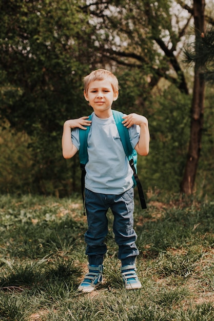 Portrait of cute blonde schoolboy in blue tshirt outdoors in spring Little boy is dissapointed by necessity to go to school Unmotivated schoolboy green park on a background