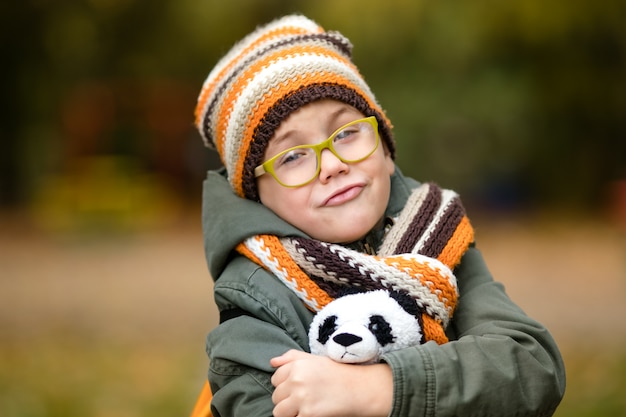 The portrait of cute boy in the glasses and warm knitting hat and scarf with his toy panda in the autumn park