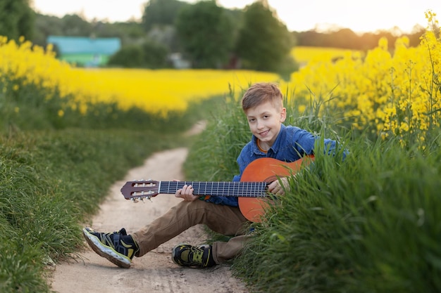 Portrait of cute boy playing a guitar sitting on road in the summer yellow field.