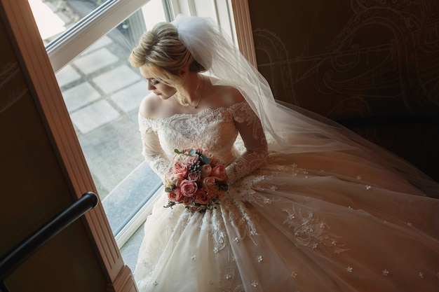 Portrait of cute bride with bridal bouquet of pink roses indoor. Pretty happy bride in luxury dress and long veil near the window. young  bride with a beautiful neckline holding bouquet of flowers.