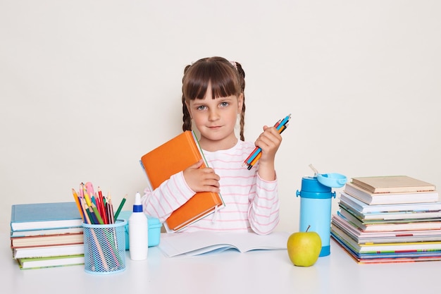 Portrait of cute charming little schoolgirl with dark hair and braids sitting at table holding book and pencils looking at camera waiting her teacher and starting lesson