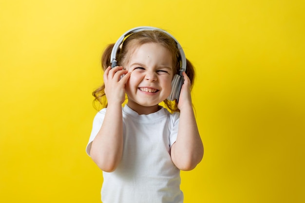 Portrait of a cute cheerful little girl listening to music with white headphones. audiobooks, audio lessons. concept of education. photo studio, yellow background, text space. High quality photo