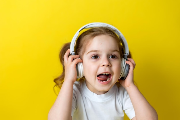 Portrait of a cute cheerful little girl listening to music with white headphones. audiobooks, audio lessons. concept of education. photo studio, yellow background, text space. High quality photo