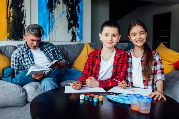 Portrait of cute girl  and boy painting with colorful gouache while  their grandfather reading his book. Family day.