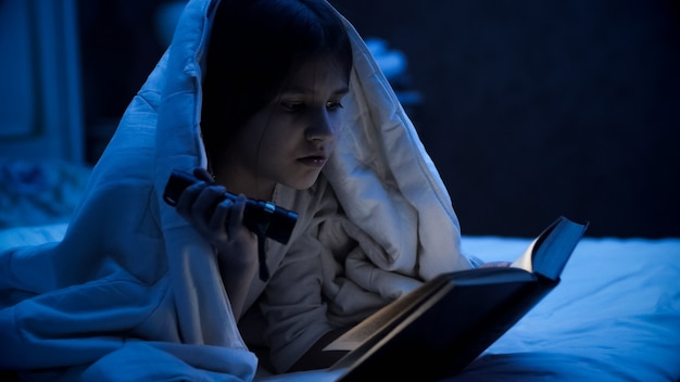 Portrait of cute girl covering with blanket and reading book with light torch.