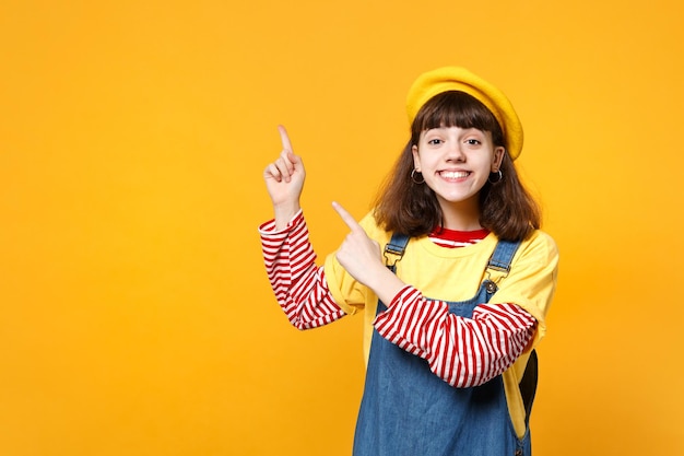 Portrait of cute girl teenager in french beret, denim sundress pointing index fingers aside up isolated on yellow background in studio. People sincere emotions, lifestyle concept. Mock up copy space.