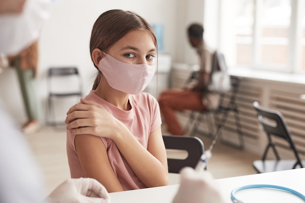 Portrait of cute girl wearing pink mask and looking at camera while waiting for vaccination in clinic, copy space