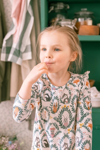 Portrait cute happy candid caucasian little kid girl five years old with blue eyes in beautiful dress with pattern animals have fun at home in kitchen helping mother to cook and licking your finger