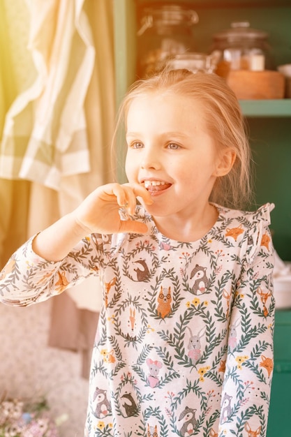 Portrait cute happy candid caucasian little kid girl five years old with blue eyes in beautiful dress with pattern animals have fun at home in kitchen helping mother to cook and licking your finger