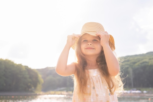 portrait of cute kid girl smiling in dress and straw hat on nature on sunny summer day