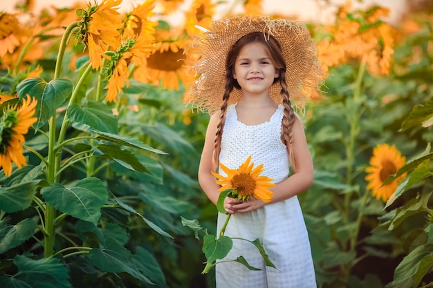 Portrait of a cute laughing girl in light clothes and a big straw hat at sunset on a field of sunflowers. she holds a big flower in her hands