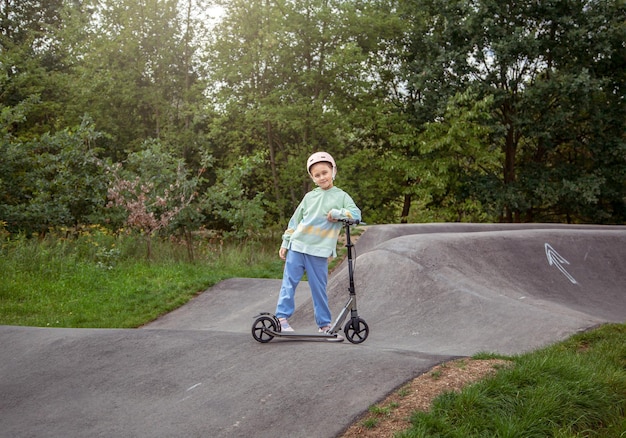 Portrait of cute little caucasian school girl wear helmet enjoy having fun riding scooter on asphalted trackxAin street park outdoors on sunny day Healthy sport children activities outsid