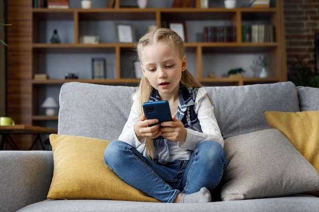 Portrait of cute little cute girl holding smartphone and resting at sofa at home Choosing favorite music or cartoonstexting messagesbrowsing internetwatching video playing games on mobile phone