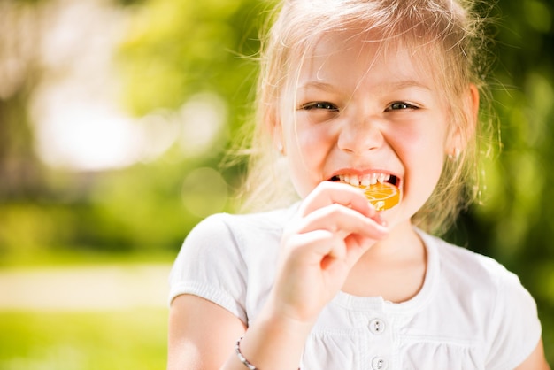 Portrait of Cute Little Girl Standing in the Park and holding in hand orange Lollipop.