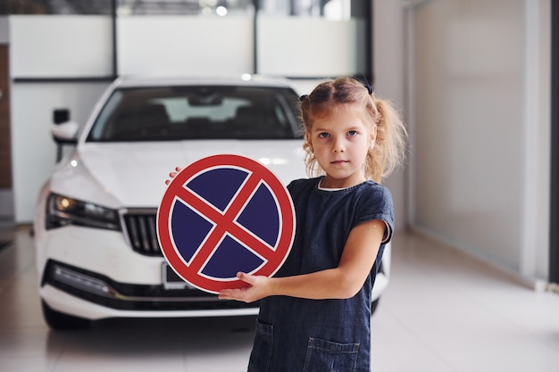 Portrait of cute little girl that holds road sign in hands in automobile salon.