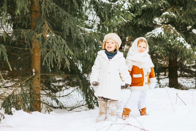 Portrait cute little girl toddler in white russian style orenburg down shawl scarf and little boy in fur coat knitted hat felt boots sit on sled in snow winter forest concept of retro Christmas