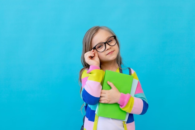 Portrait of a cute little girl with glasses in a striped jacket with notebooks and a backpack adjusts her glasses like a teacher Concept of education Photo studio blue background place for text