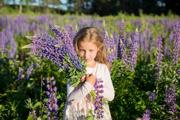 Portrait of cute little happy seven year old kid girl with bloom flowers lupines in field of purple flowers Child in nature concept Summer vacation holidays Spring allergy season Childhood