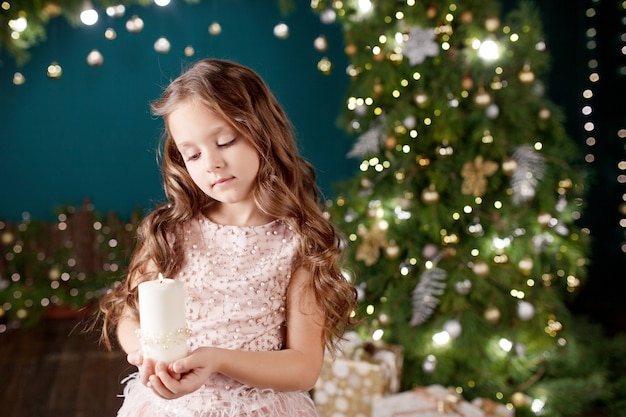 Portrait of cute long-haired little girl in dress on  of  lights. Little girl holding burning candle. Christmas, New Year .