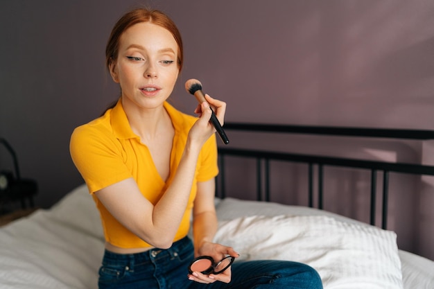 Photo portrait of cute redhead young woman applying nude facial powder to get natural effect with big professional makeup brush sitting on bed in bedroom