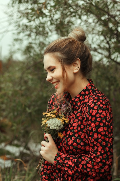 Portrait of a cute smiling girl in a dress with red flowers, which holds a bouquet of wildflowers in her hands.