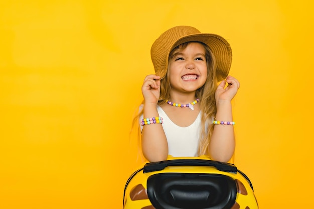 Portrait of a cute smiling girl in a straw hat with a suitcase studio photo on a yellow background