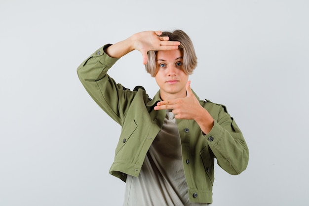 Portrait of cute teen boy showing frame gesture with finger guns in green jacket and looking confident front view