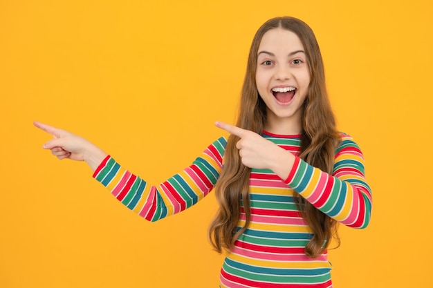 Portrait of cute teenager child girl pointing hand showing adverts with copy space over yellow background Excited face cheerful emotions of teenager girl