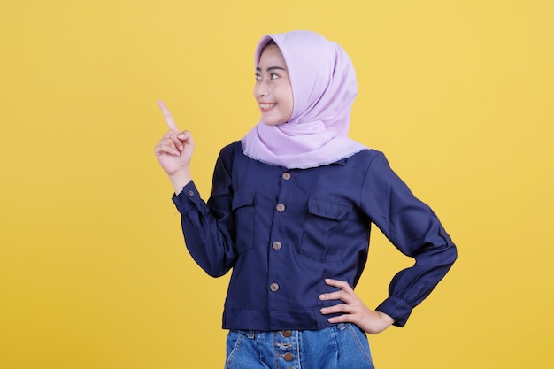 Portrait of cute woman pointing up in casual cloth wearing hijab on isolated yellow background