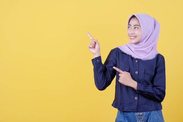 Portrait of cute woman pointing up in casual cloth wearing hijab on isolated yellow background