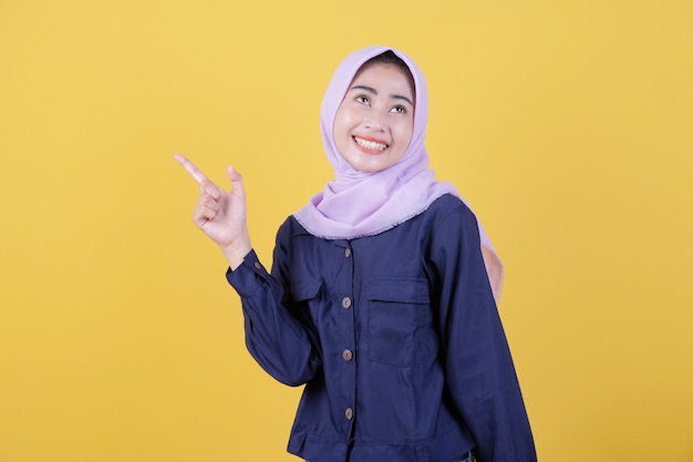 Portrait of cute woman pointing up in casual cloth wearing hijab on isolated yellow background