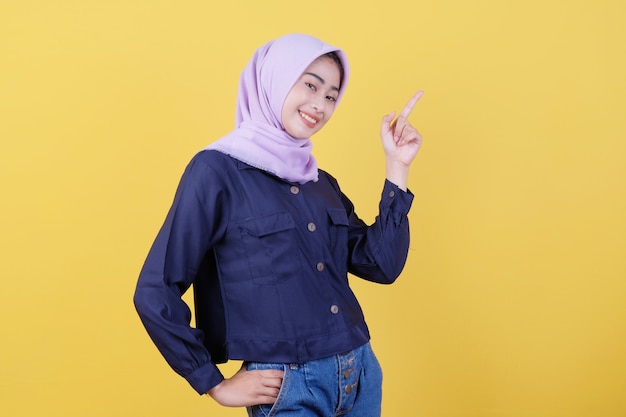 Portrait of cute woman pointing up in casual cloth wearing hijab on isolated yellow background