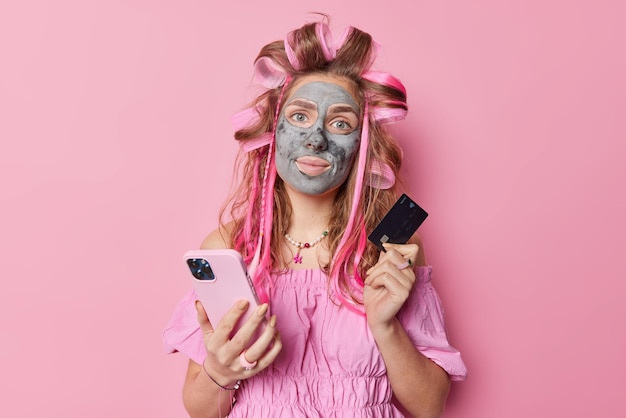 Portrait of cute woman thinks of making shopping online holds smartphone and plastic credit card applies hair curlers clay mask for skin rejuvenation wears blouse isolated over pink background