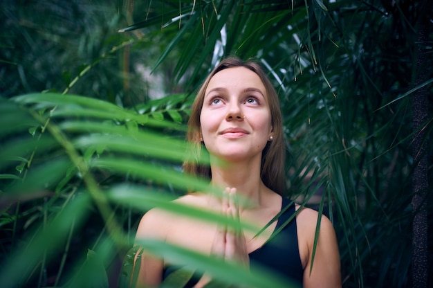 Portrait of cute young woman meditating and doing namaste hand in jungle.