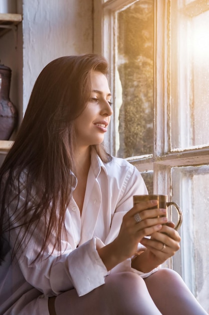 Portrait cute young woman in white shirt drinking tea and looking through window while sitting at windowsill indoors