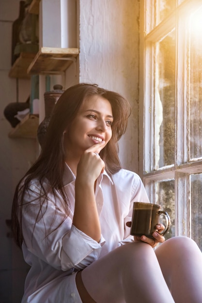 Portrait cute young woman in white shirt drinking tea and looking through window while sitting at windowsill indoors