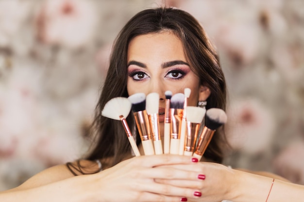 A portrait of a cute young woman with beautiful eyes, who is holding makeup brushes and representing a nice makeup on her face.