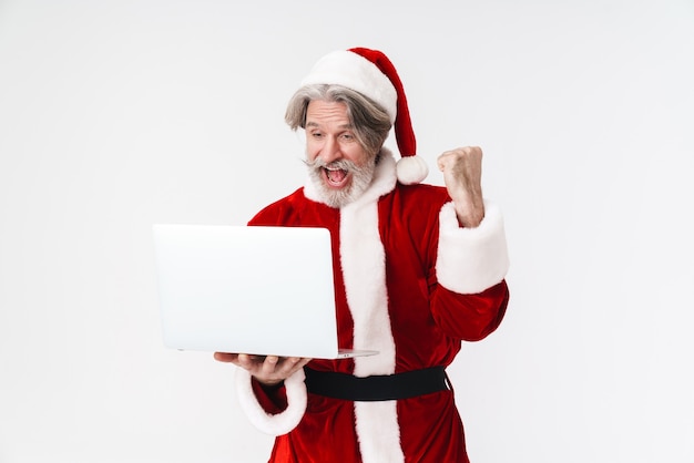 Portrait of delighted old man wearing Santa Claus red costume making winner gesture and using laptop isolated on white