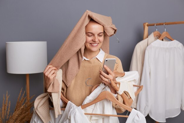 Portrait of delighted satisfied woman shopper with clothes on her head holding clothing on hangers standing with smartphone in hands looking at display with toothy smile