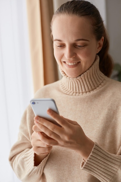 Photo portrait of delighted young adult woman with ponytail hairstyle wearing beige casual style sweater using mobile phone, writing post for social networks or watching photos.