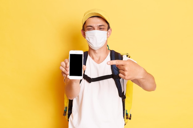 Portrait of delivery man wearing protective mask holding thermo bag and showing screen of phone with empty space for advertisement isolated over yellow background