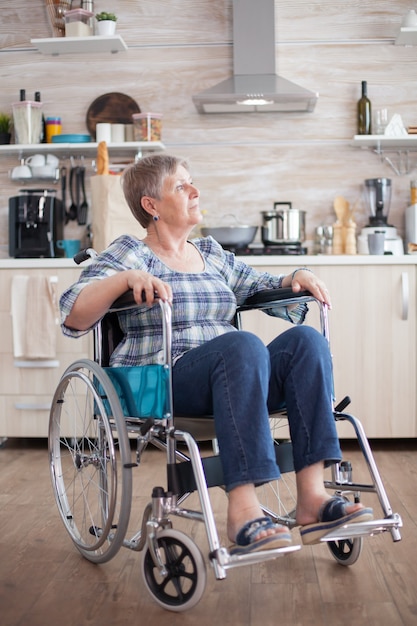 Portrait of disabled unhappy senior woman in wheelchair looking lost in kitchen. Elderly handicapped pensioner after injury and rehab, paralysis and disability for depressed invalid full of sorrow, wo