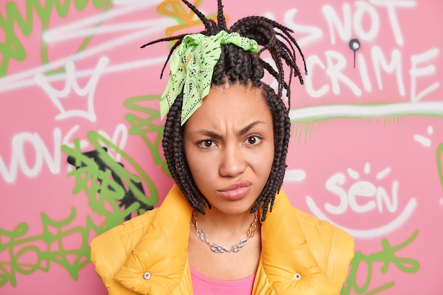 Portrait of displeased woman with combed dreadlocks dressed in stylish clothes poses against colorful graffiti wall belongs to youth subculture