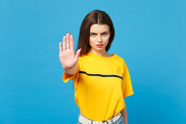 Portrait of displeased young woman in vivid casual clothes looking camera, showing stop gesture with palm isolated on bright blue background in studio. People lifestyle concept. Mock up copy space.