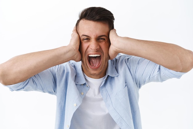 Portrait of distressed and angry young man going insane shut ears and shouting displeased feel depressed and unstable having mental breakdown or emotional burnout white background