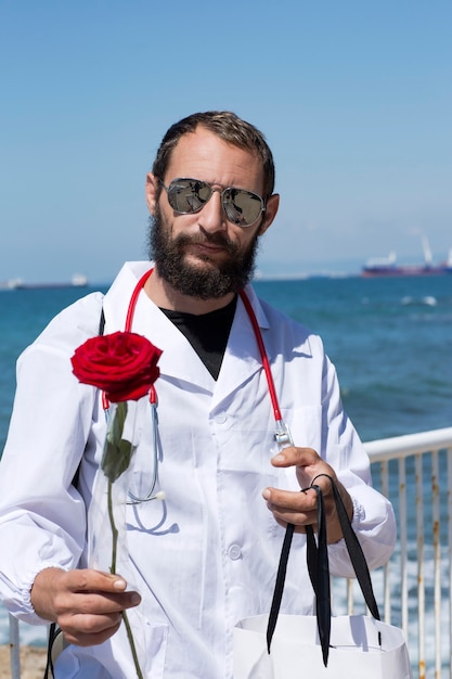 Portrait of doctor in a white coat with stethoscope and sunglasses holding a red rose flower in hand. American handsome bearded man on blue sky background. The concept of recovery, gratitude to doctor