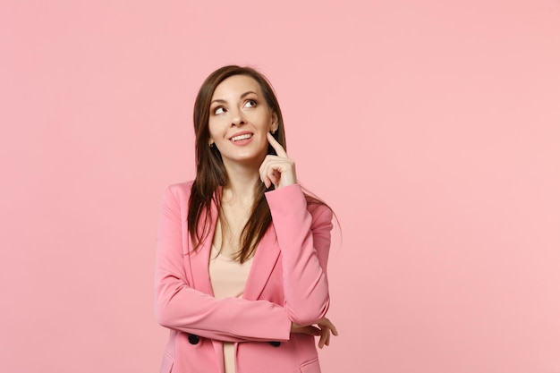 Portrait of dreamful pretty young woman in jacket looking up put hand prop up on chin isolated on pastel pink wall background in studio. People sincere emotions, lifestyle concept. Mock up copy space.