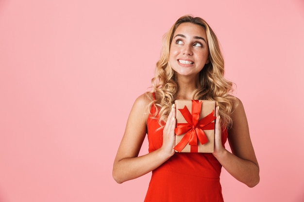 Portrait of a dreaming pretty excited happy young cute blonde woman posing isolated over pink wall holding present gift box.