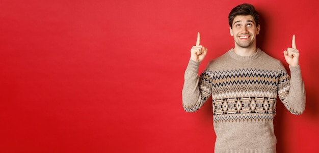 Portrait of dreamy handsome guy thinking about christmas holidays wearing winter sweater pointing and looking up at new year promo offer standing over red background